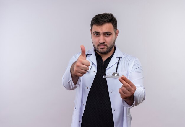 Jovem médico barbudo vestindo jaleco branco com estetoscópio segurando o termômetro e olhando para a câmera mostrando os polegares para cima com uma cara feliz