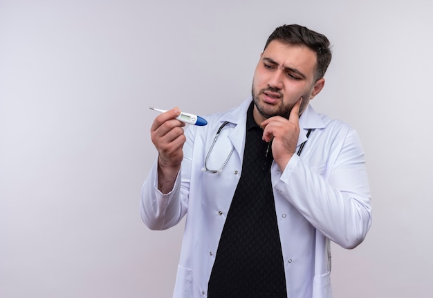 Jovem médico barbudo vestindo jaleco branco com estetoscópio segurando um termômetro digital olhando para ele com uma expressão pensativa no rosto