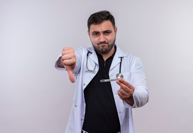 Jovem médico barbudo vestindo jaleco branco com estetoscópio segurando um termômetro e mostrando antipatia com rosto infeliz