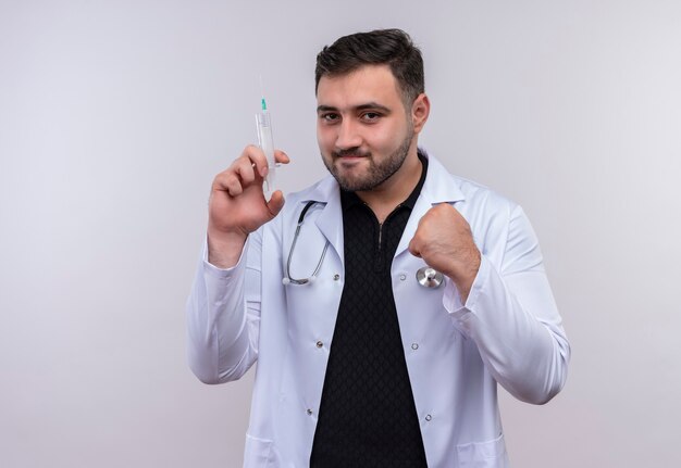 Jovem médico barbudo vestindo jaleco branco com estetoscópio segurando uma seringa e sorrindo confiante com o punho cerrado, conceito vencedor