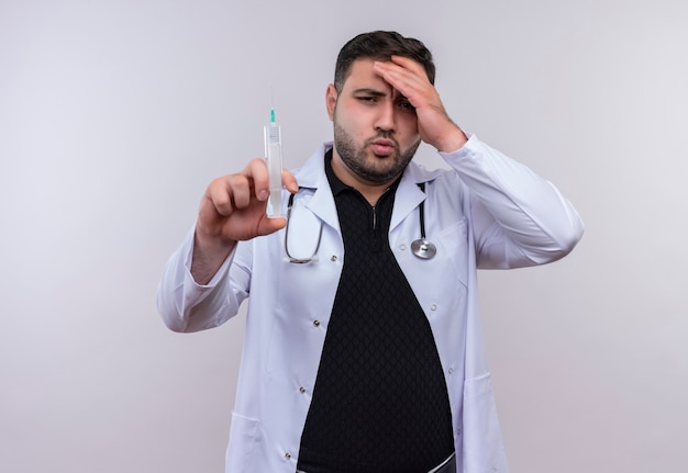 Jovem médico barbudo vestindo jaleco branco com estetoscópio segurando uma seringa, parecendo confuso e muito ansioso