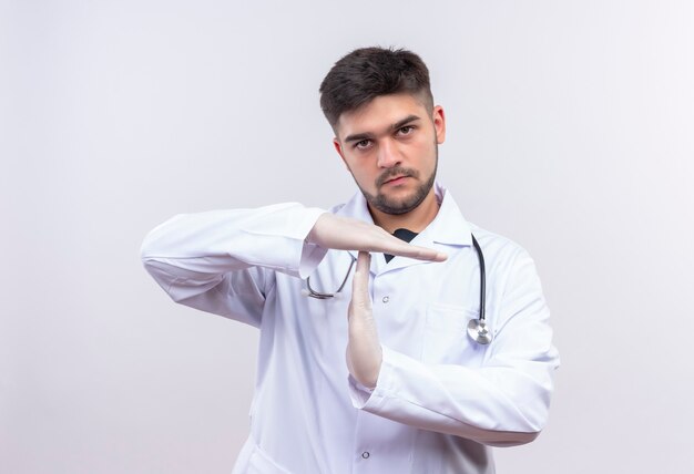 Jovem médico bonito usando jaleco branco, luvas médicas brancas e estetoscópio mostrando o intervalo com as mãos em pé sobre a parede branca