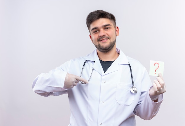 Jovem médico bonito usando jaleco branco, luvas médicas brancas e estetoscópio, sorrindo, apontando para a pergunta, sinalizar em pé sobre uma parede branca