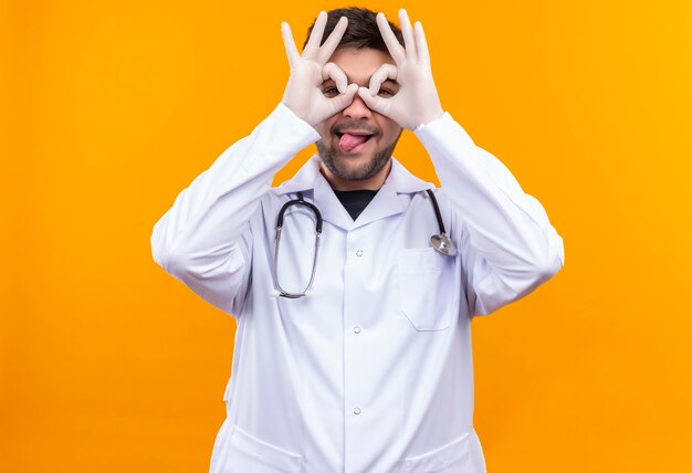 Foto grátis jovem médico bonito usando jaleco branco, luvas médicas e estetoscópio, fechando os olhos de brincadeira, fazendo óculos em pé sobre a parede laranja