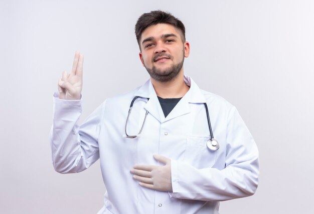 Jovem médico bonito usando jaleco branco, luvas médicas e estetoscópio segurando a barriga, mostrando o símbolo da paz em pé sobre uma parede branca