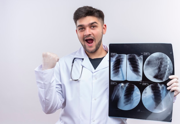 Foto grátis jovem médico bonito usando um avental médico branco, luvas médicas brancas e estetoscópio, feliz com os resultados da tomografia em pé sobre uma parede branca