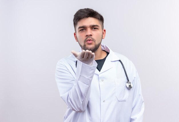 Jovem médico bonito usando um avental médico branco, luvas médicas brancas e estetoscópio mandando beijos em pé sobre uma parede branca