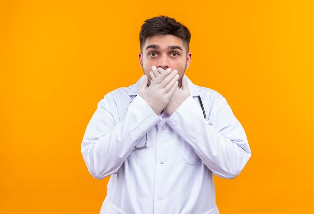 Jovem médico bonito usando um avental médico branco, luvas médicas e estetoscópio com medo de fechar o mouse com as mãos em pé sobre a parede laranja