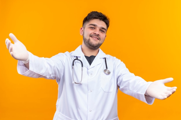 Jovem médico bonito vestindo jaleco branco com luvas médicas e estetoscópio dando as boas-vindas em pé sobre a parede laranja