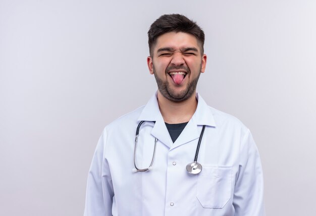 Jovem médico bonito vestindo jaleco branco e estetoscópio mostrando a língua em pé sobre uma parede branca de maneira divertida