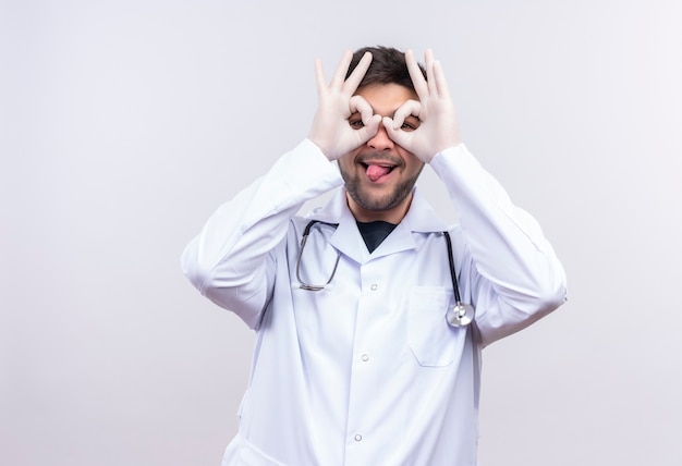 Jovem médico bonito vestindo jaleco branco, luvas médicas brancas e estetoscópio, fechando os olhos de forma divertida com a mão mostrando a língua em pé sobre uma parede branca