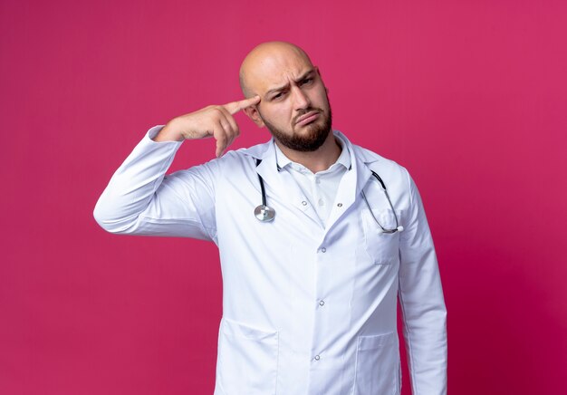 Jovem médico careca confuso, vestindo bata médica e estetoscópio colocando o dedo na testa, isolado na parede rosa