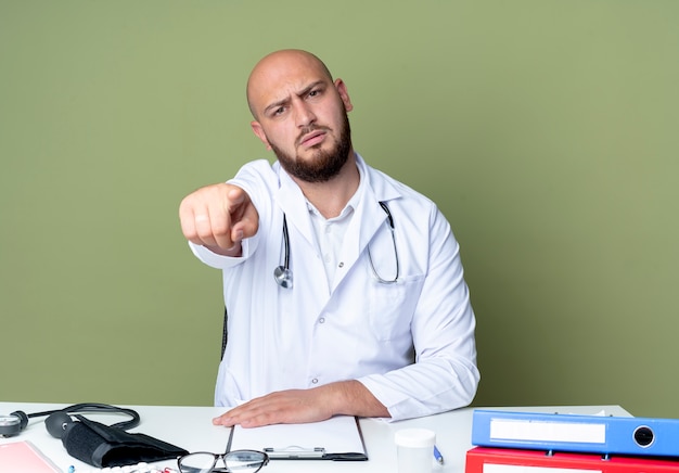 Jovem médico careca rigoroso usando túnica médica e estetoscópio sentado na mesa trabalhando com ferramentas médicas, mostrando seu gesto isolado na parede verde