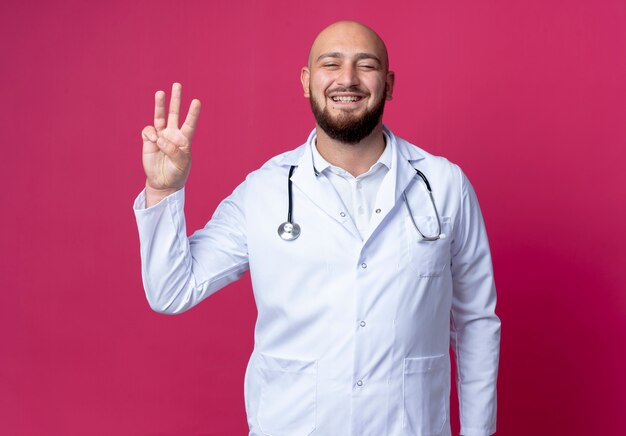 Jovem médico careca satisfeito, vestindo bata médica e estetoscópio mostrando três isolados na parede rosa