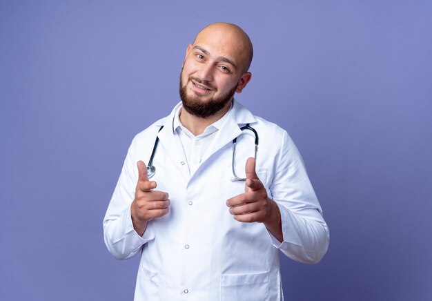 Jovem médico careca satisfeito, vestindo bata médica e estetoscópio, mostrando um gesto isolado sobre fundo azul