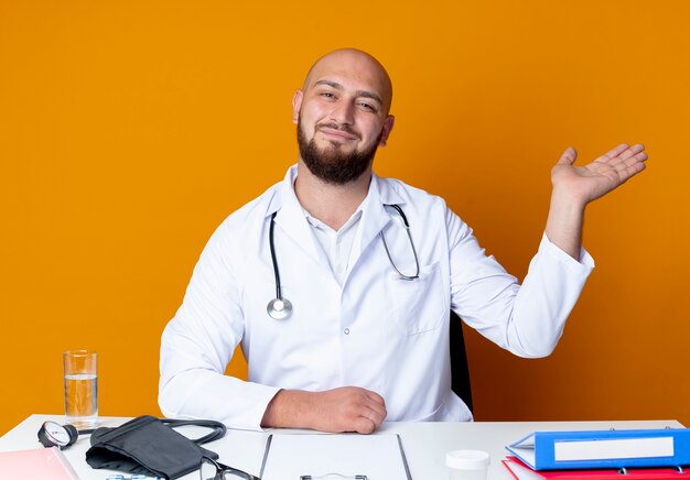 Jovem médico careca satisfeito, vestindo túnica médica e estetoscópio sentado na mesa de trabalho com pontos de ferramentas médicas com a mão ao lado isolada em fundo laranja