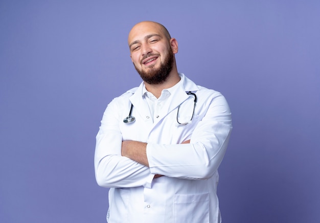 Jovem médico careca sorridente, vestindo túnica médica e estetoscópio cruzando as mãos isoladas sobre fundo azul