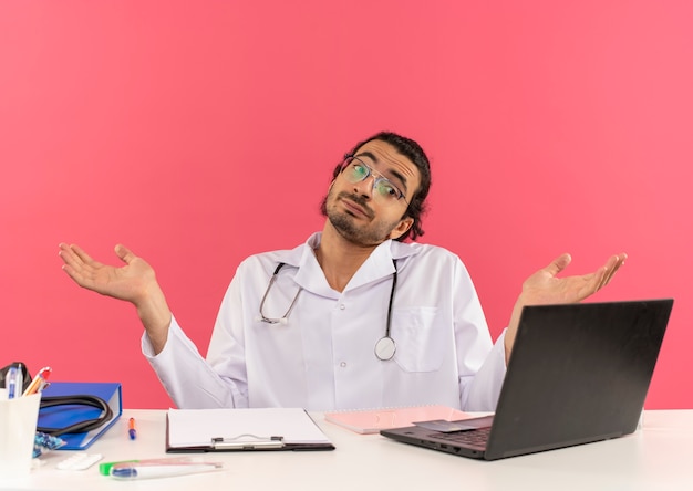 Jovem médico confuso com óculos de médico, vestindo túnica médica com estetoscópio sentado na mesa, trabalhando no laptop com ferramentas médicas, espalhando as mãos no fundo rosa isolado com espaço de cópia
