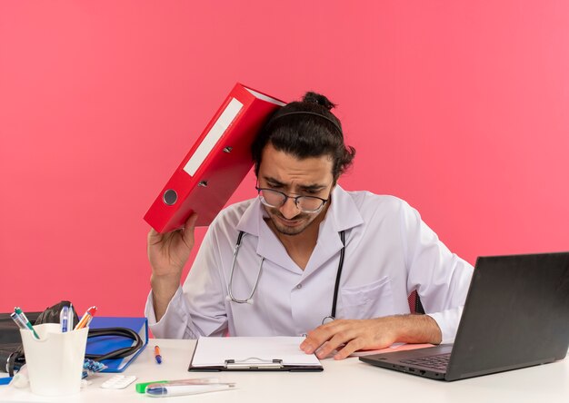 Jovem médico confuso com óculos médicos, usando roupão médico e estetoscópio sentado na mesa