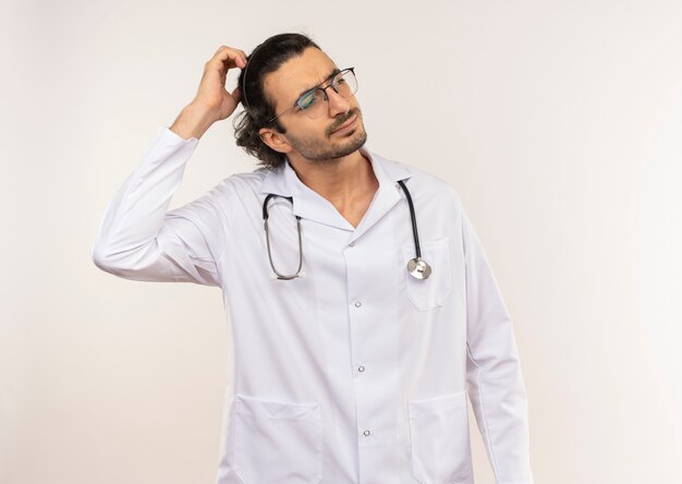 Jovem médico confuso com óculos ópticos, manto branco e estetoscópio colocando a mão na cabeça