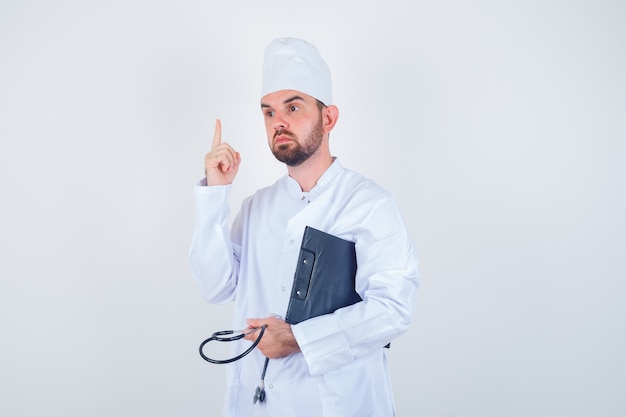 Jovem médico de uniforme branco segurando a prancheta, estetoscópio, apontando para cima e olhando a vista frontal inteligente.