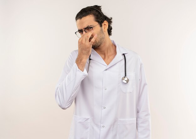 Jovem médico enjoado com óculos ópticos, túnica branca e estetoscópio com o nariz fechado na parede branca isolada com espaço de cópia