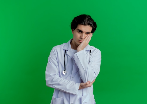 Foto grátis jovem médico entediado usando roupão médico e estetoscópio, segurando o rosto e parecendo isolado