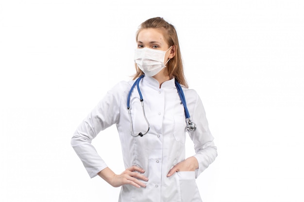 jovem médico feminino no terno médico branco no estetoscópio de máscara protetora branco posando no branco