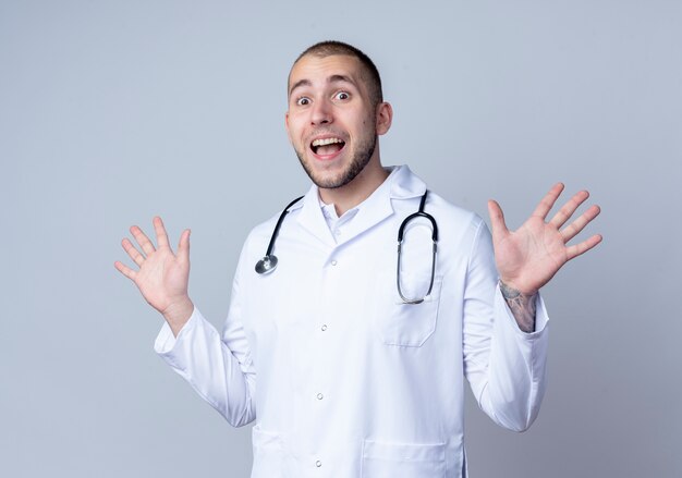 Jovem médico impressionado usando túnica médica e estetoscópio no pescoço, mostrando as mãos vazias isoladas no branco