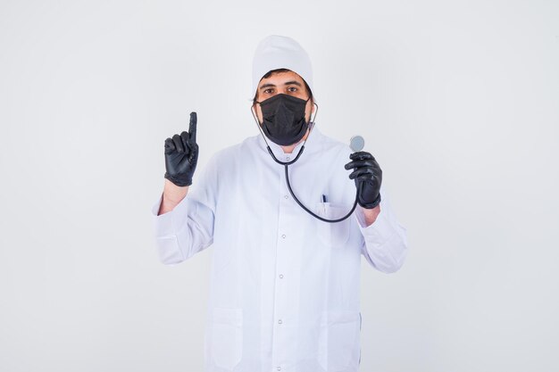 Jovem médico masculino apontando para cima enquanto segura o estetoscópio em uniforme branco e parece confiante, vista frontal.