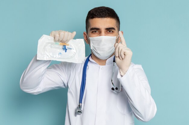 Jovem médico masculino de terno branco com estetoscópio azul segurando um pacote de injeção