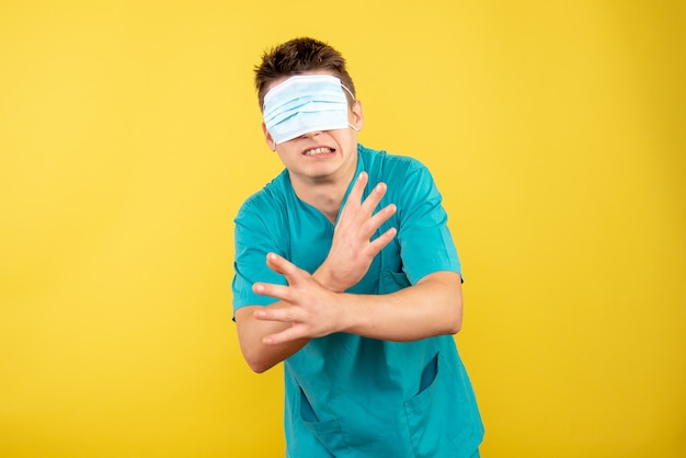 Jovem médico masculino em terno médico com máscara sobre os olhos e fundo amarelo.