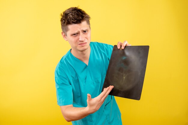 Jovem médico masculino em terno médico segurando um raio-x em fundo amarelo de frente