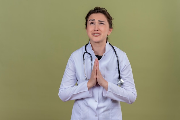 Jovem médico preocupado, usando bata médica e estetoscópio, mostrando gesto de oração na parede verde