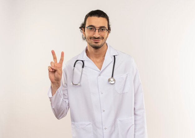 Jovem médico satisfeito com óculos ópticos, túnica branca e estetoscópio, mostrando um gesto de paz em uma parede branca isolada com espaço de cópia