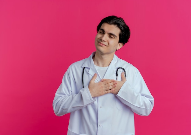 Foto grátis jovem médico satisfeito, vestindo bata médica e estetoscópio, mantendo as mãos no coração com os olhos fechados, isolado na parede rosa com espaço de cópia