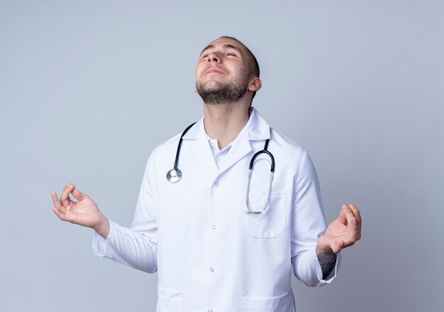 Jovem médico satisfeito, vestindo túnica médica e estetoscópio em volta do pescoço, meditando com os olhos fechados, isolado na parede branca