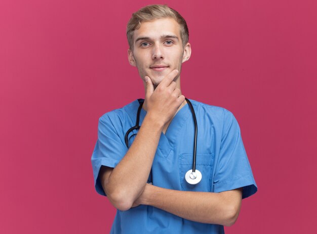 Jovem médico satisfeito vestindo uniforme de médico com estetoscópio segurando o queixo isolado na parede rosa