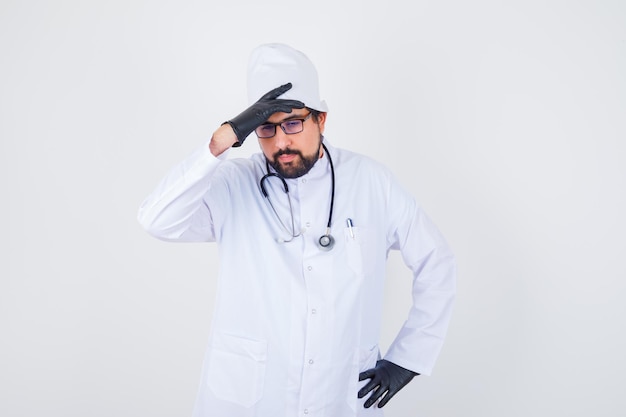 Jovem médico segurando a mão na testa em uniforme branco e parecendo exausto, vista frontal.