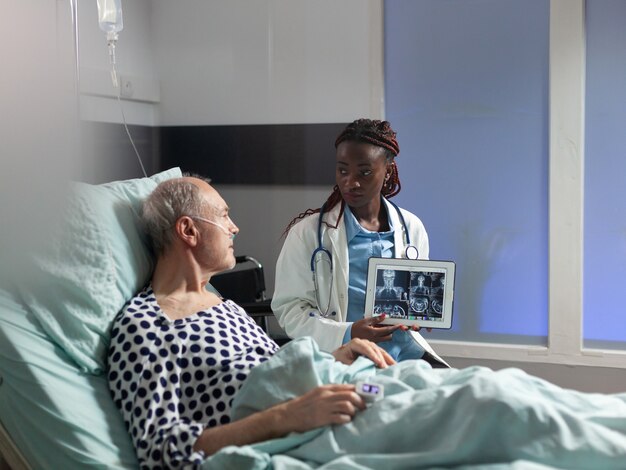 Jovem médico sentado ao lado de um homem sênior, explicando o diagnóstico de trauma corporal, mostrando um raio-x no tablet pc em um quarto de hospital