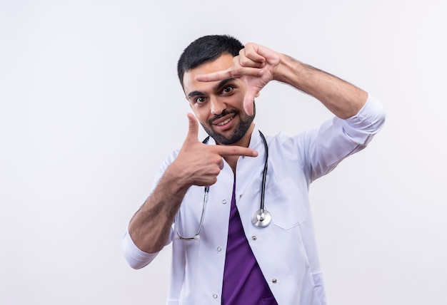 Jovem médico sorridente com bata médica de estetoscópio mostrando gesto de foto em fundo branco isolado