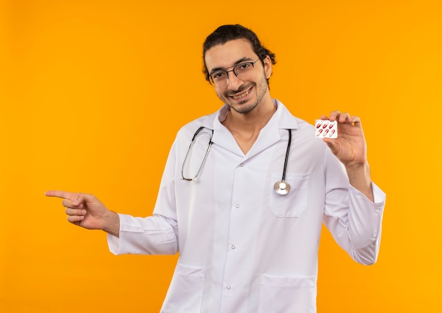 Jovem médico sorridente com óculos médicos, usando roupão médico, com um estetoscópio segurando os comprimidos e apontando para os lados em amarelo