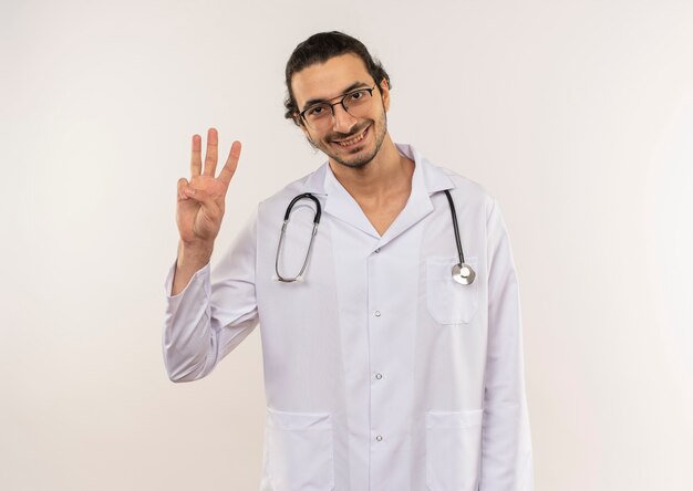 Jovem médico sorridente com óculos ópticos, túnica branca e estetoscópio mostrando três em uma parede branca isolada com espaço de cópia