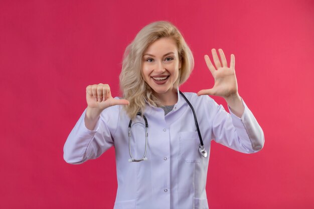 Jovem médico sorridente usando estetoscópio com bata médica, mostrando um gesto diferente no fundo vermelho