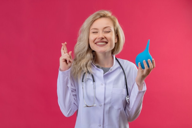 Jovem médico sorridente, usando estetoscópio e bata médica, segurando os dedos cruzados em um backgroung vermelho