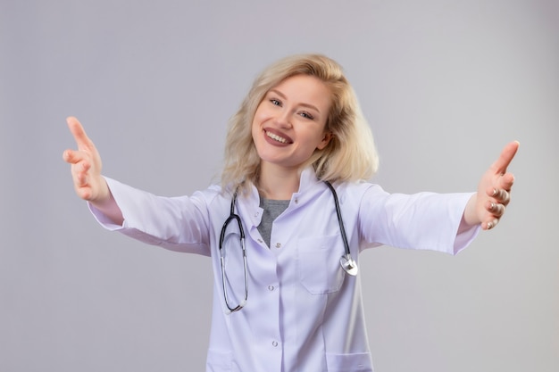 Jovem médico sorridente usando estetoscópio e vestido de médico, mostrando um gesto de abraço na parede branca