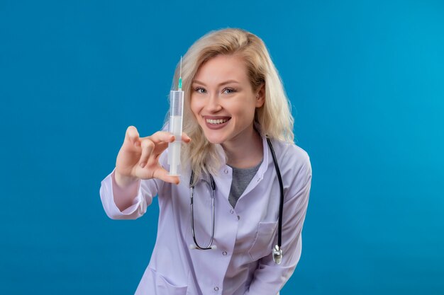 Jovem médico sorridente usando estetoscópio e vestido de médico segurando uma seringa na parede azul