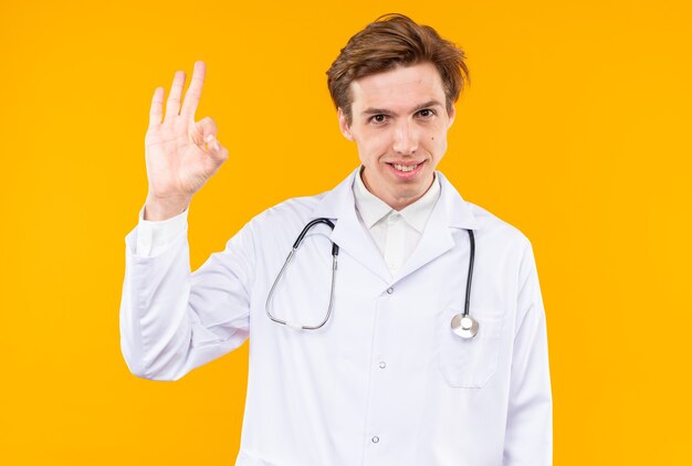 Jovem médico sorridente usando manto médico com estetoscópio mostrando um gesto de aprovação