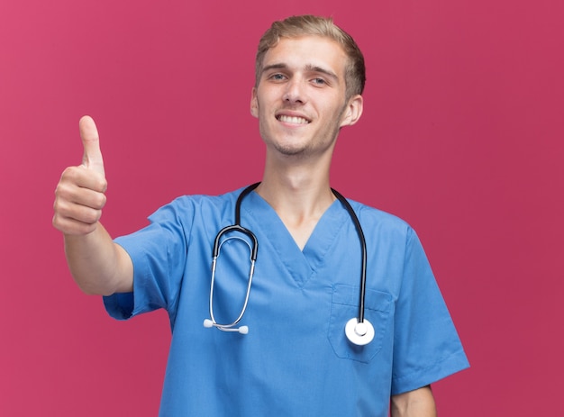 Jovem médico sorridente usando uniforme de médico com estetoscópio aparecendo o polegar isolado na parede rosa