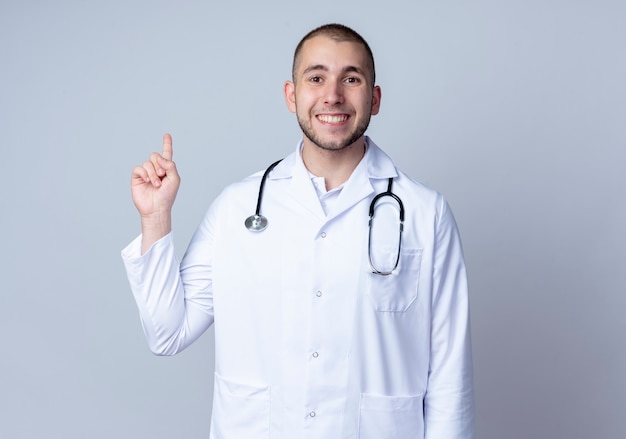 Jovem médico sorridente, vestindo bata médica e estetoscópio em volta do pescoço, levantando o dedo isolado na parede branca